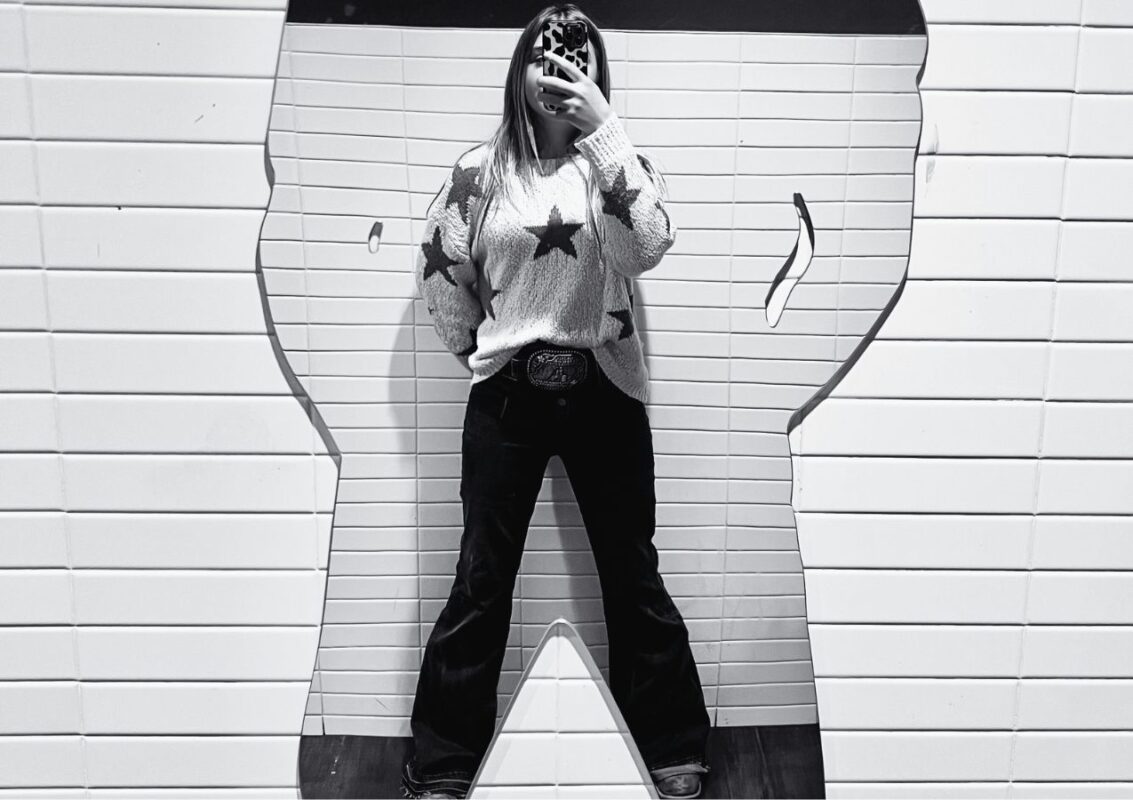 Image of young woman taking picture of self in mirror shaped like a large cowboy_ Mirror poem by Dawn Francis_Rebel-Faith_2024